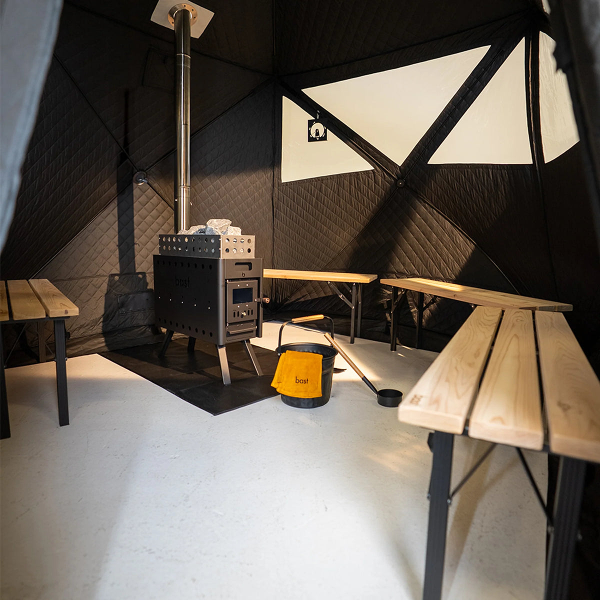 Wood-burning stove in a Bast Sauna tent for 4-10 people with geometric wall design and wooden benches, and a saunta bucket and ladel.