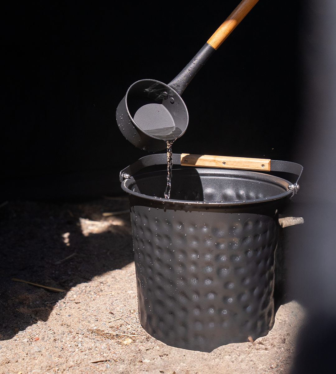 Black bucket with a wooden handle being used to pour water in a BAST sauna tent.