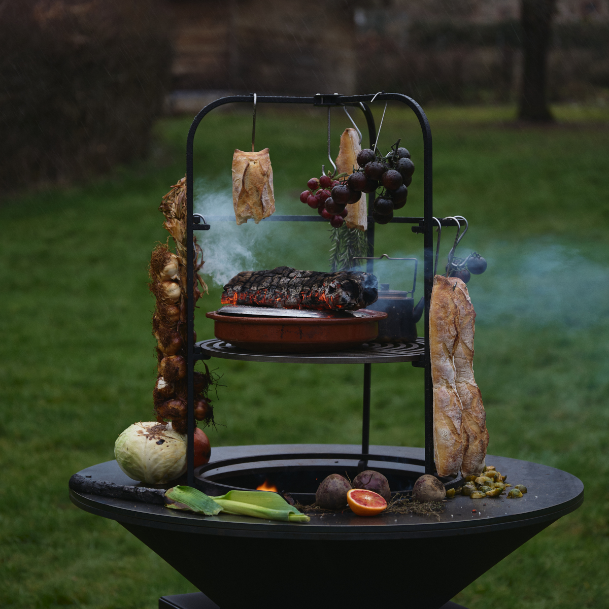 OFYR Cage 100 with bread, tomatoes and herbs hanging above the fire and vegetables on the cooking plate.