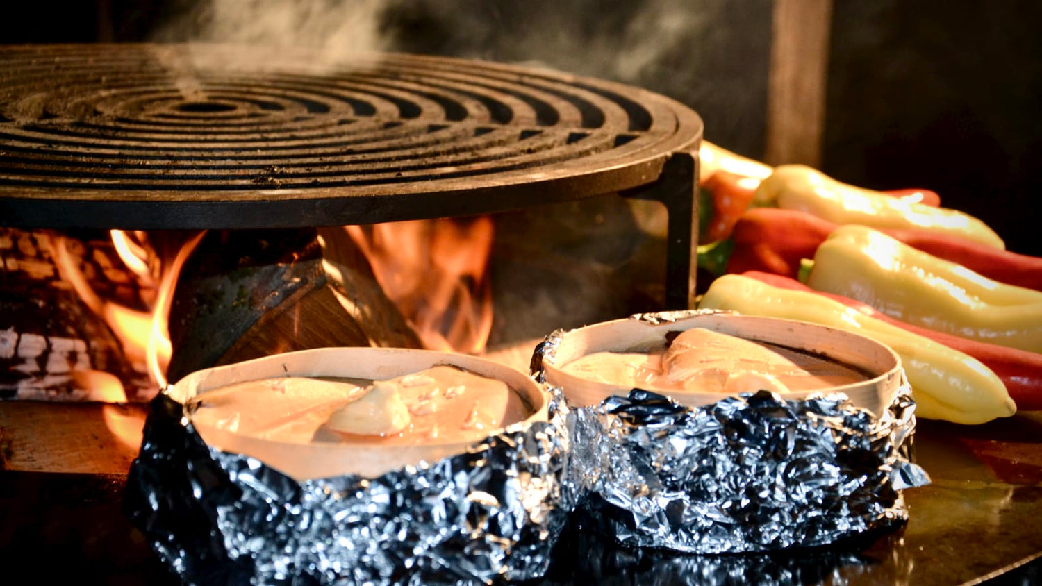 Vacherin Mont d’Or fondue in its traditional wooden box on the OFYR grill, with colorful bell peppers on the side and open fire in the background.