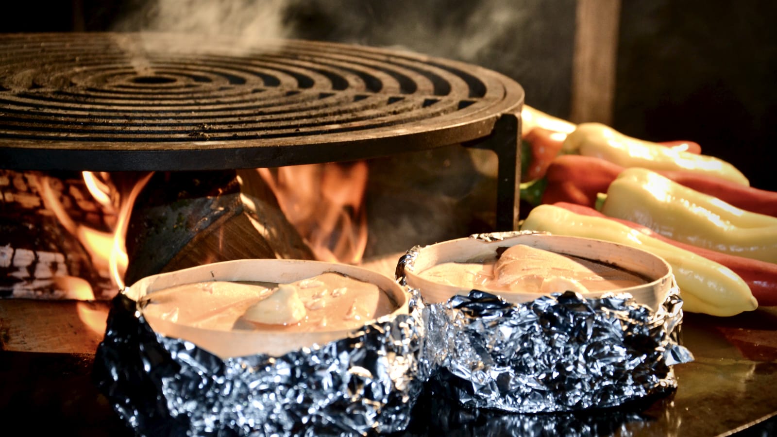 Vacherin Mont d’Or fondue in its traditional wooden box on the OFYR grill, with colorful bell peppers on the side and open fire in the background.