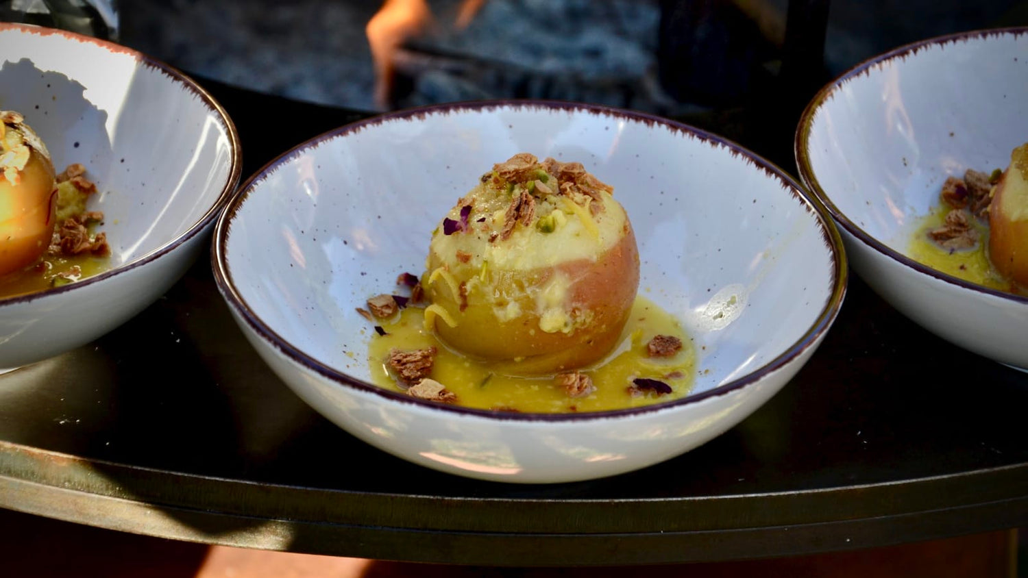 Baked apple with spiced sauce and crunchy topping, served in a white bowl on the OFYR grill – a warm and flavourful autumn dessert outdoors.