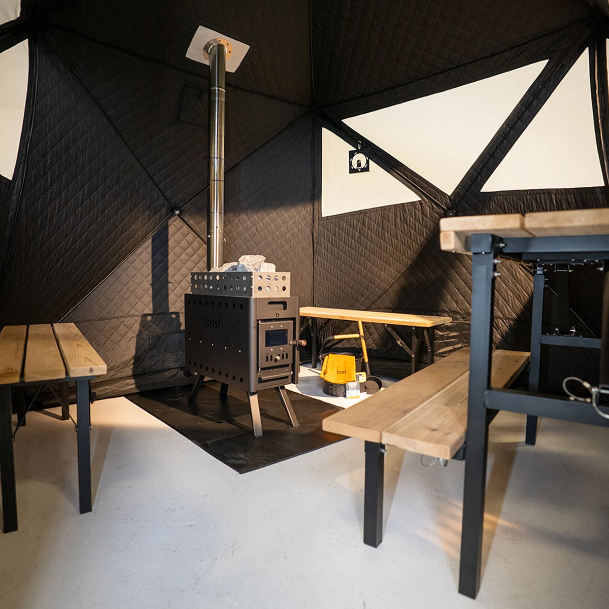 Wood-burning stove in a Bast Sauna tent (4-10 people) with geometric wall design and wooden benches, and a saunta bucket and ladel.