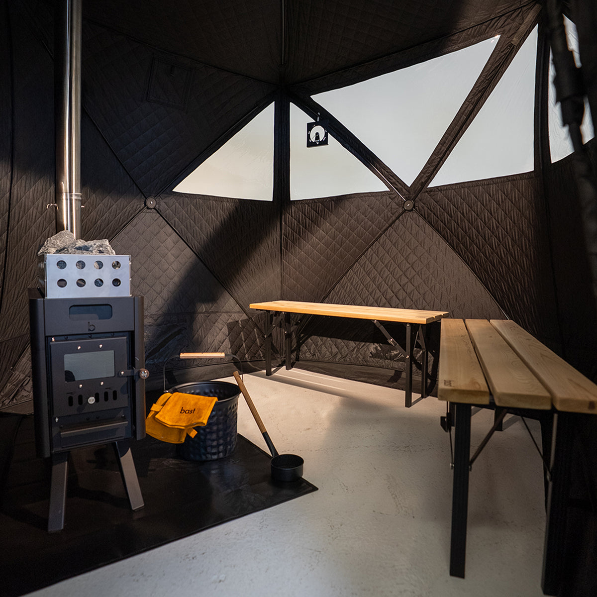 Wood-burning stove in a Bast Sauna tent  (2-4 people) with geometric wall design and wooden benches, and a saunta bucket and ladel.