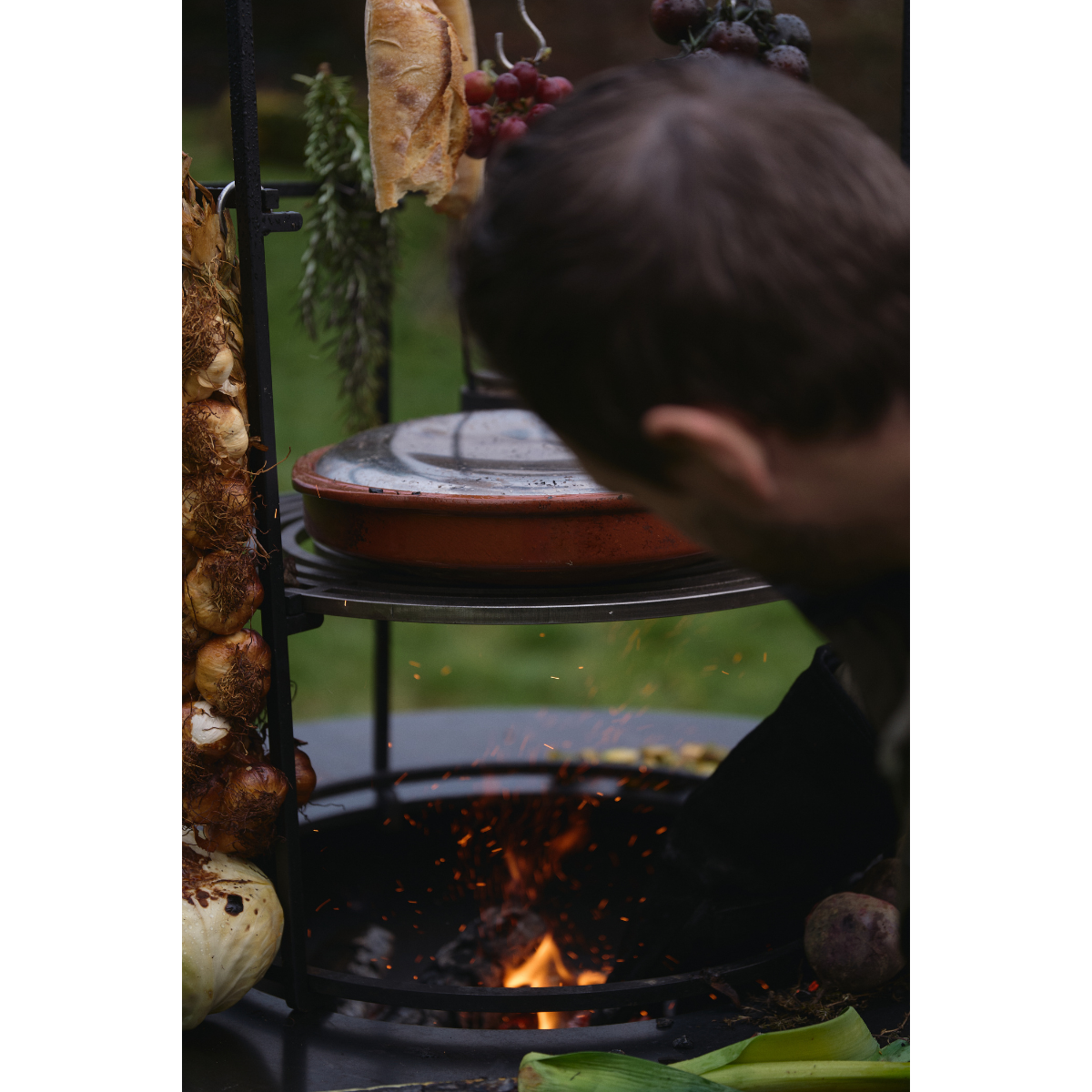 Cook tending an OFYR Cage 100 with hanging bread and tomatoes and a covered dish on the raised grill grate.