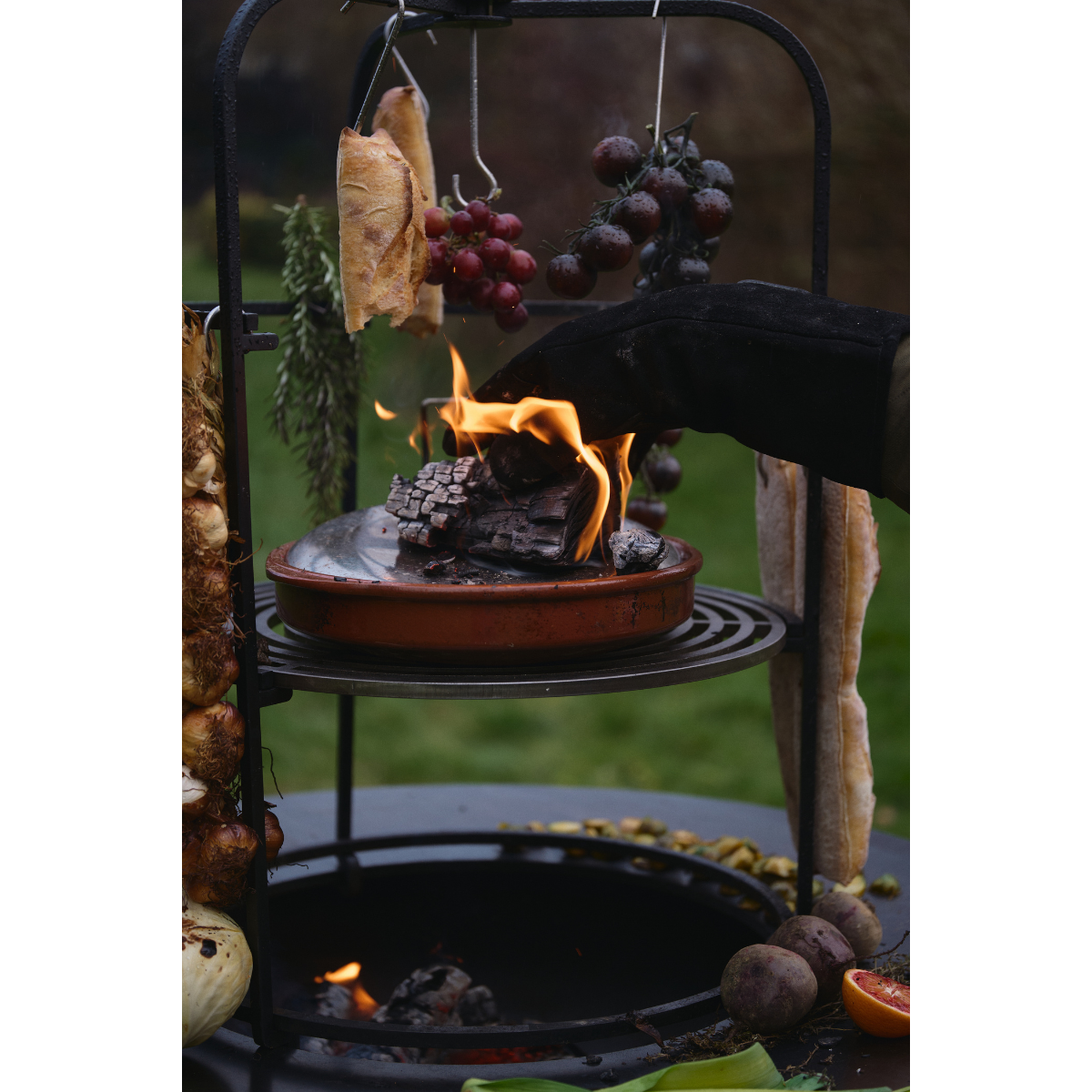 Gloved hand placing burning wood on a covered dish on the OFYR Cage 100 grill grate, beneath hanging bread and tomatoes.